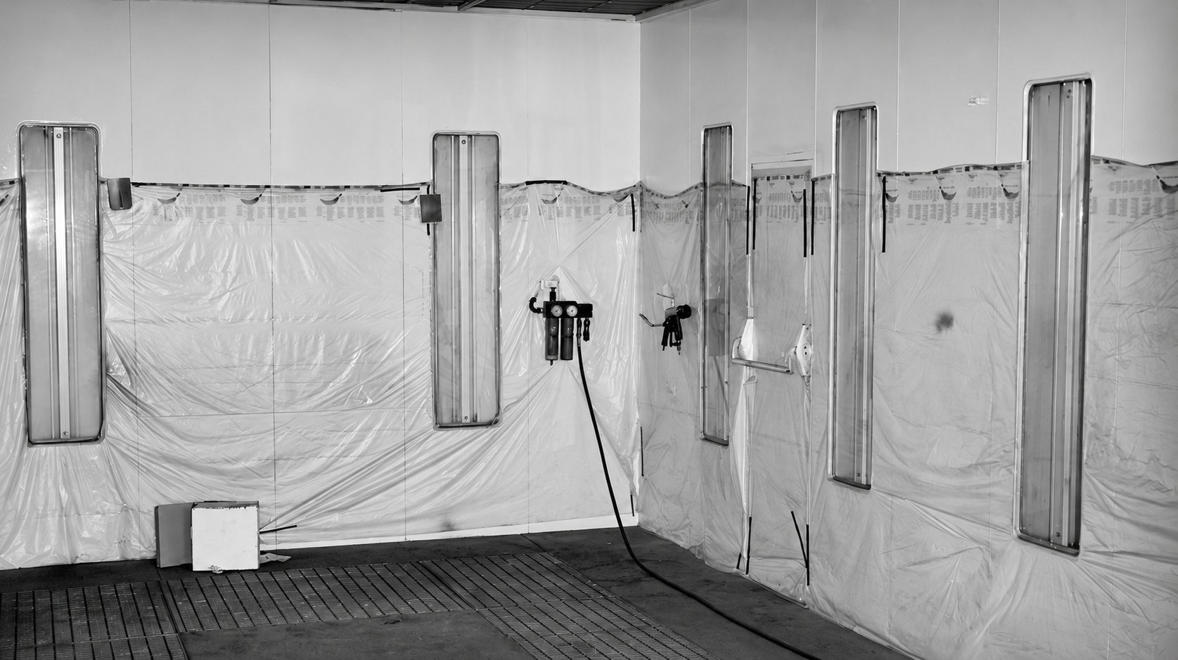Intérieur d'une cabine de peinture en noir et blanc dont les murs sont recouverts de film plastique, avec des panneaux lumineux verticaux et un détendeur d'air comprimé.