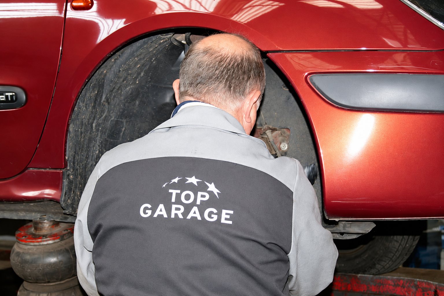 Un mécanicien en veste grise Top Garage, vu de dos, examine le passage de roue et le frein d'une voiture rouge surélevée.