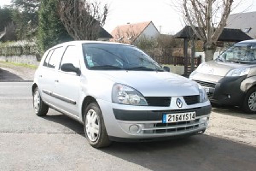 Une Renault Clio grise est garée sur un parking en extérieur, avec des arbres et des maisons à l'arrière-plan et une autre voiture sur la droite.