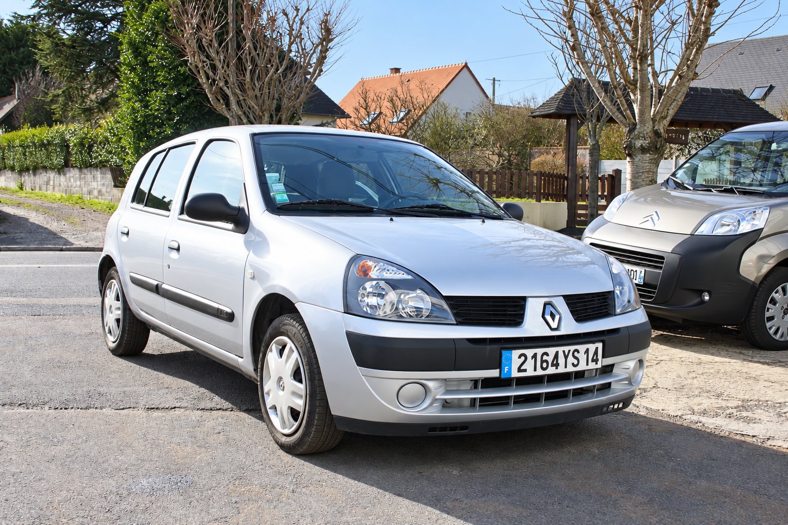 Une Renault Clio grise à cinq portes est garée dans une rue de quartier pavillonnaire, avec un autre véhicule à droite et des maisons en arrière-plan.