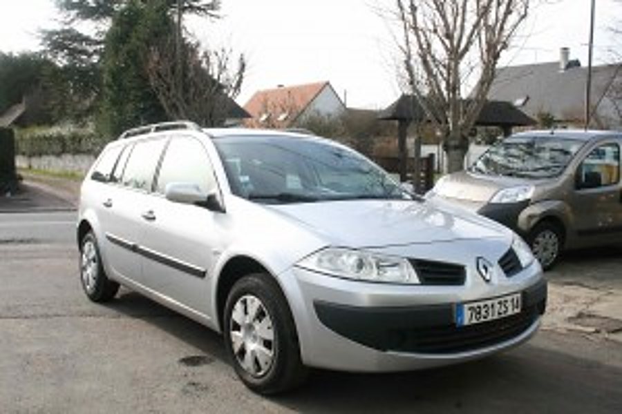Une Renault Mégane break gris argenté est garée dans une rue résidentielle, avec des maisons et des arbres en arrière-plan.