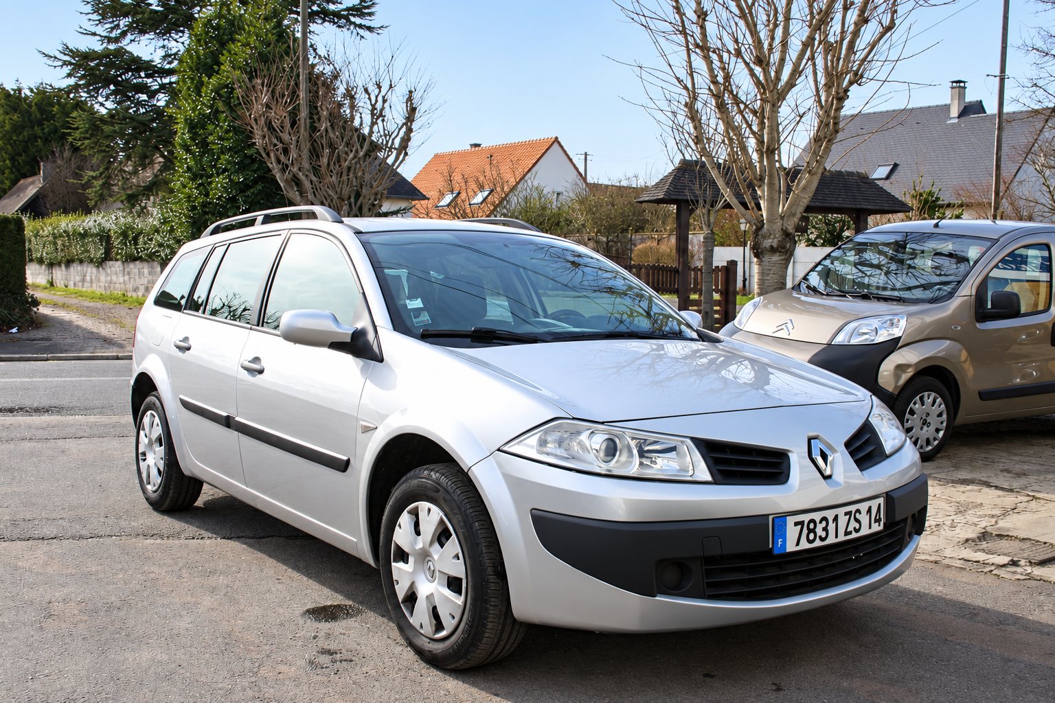 Une Renault Mégane break grise stationnée dans une rue de quartier pavillonnaire, avec un autre véhicule beige et des maisons en arrière-plan.