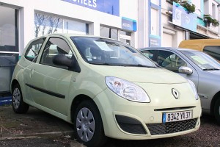 Une petite Renault Twingo jaune pâle est garée devant la façade d'un garage, avec une autre voiture grise à côté.