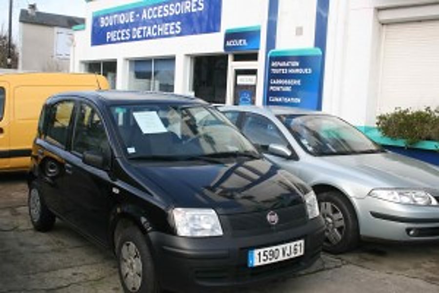Une Fiat Panda noire et une berline grise sont garées devant la façade d'un garage dont l'enseigne annonce boutique, accessoires et pièces détachées.